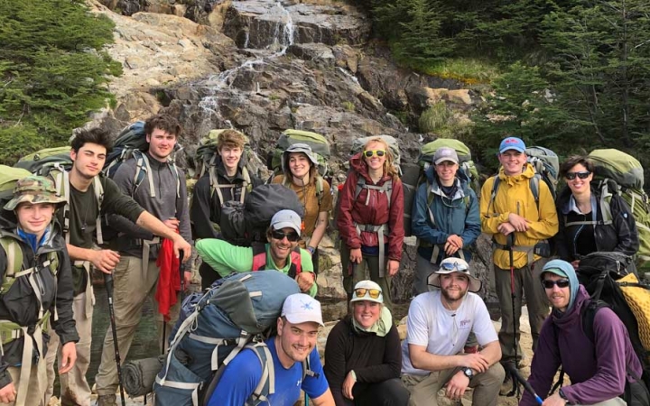 A group of people wearing backpacks pose for a photo in front of a small waterfall.